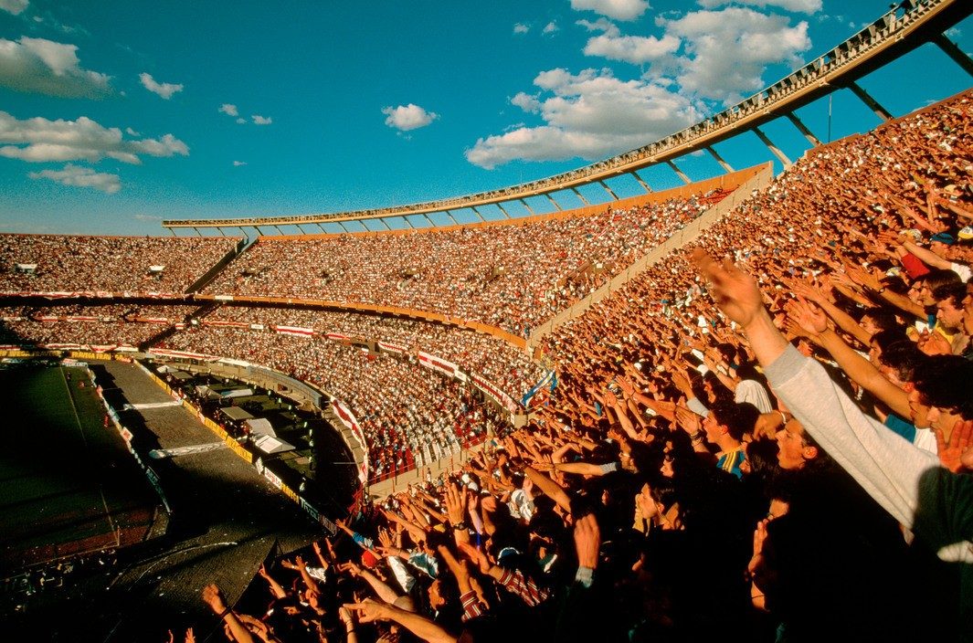 A sunlit stadium crowd on its feet, arms raised